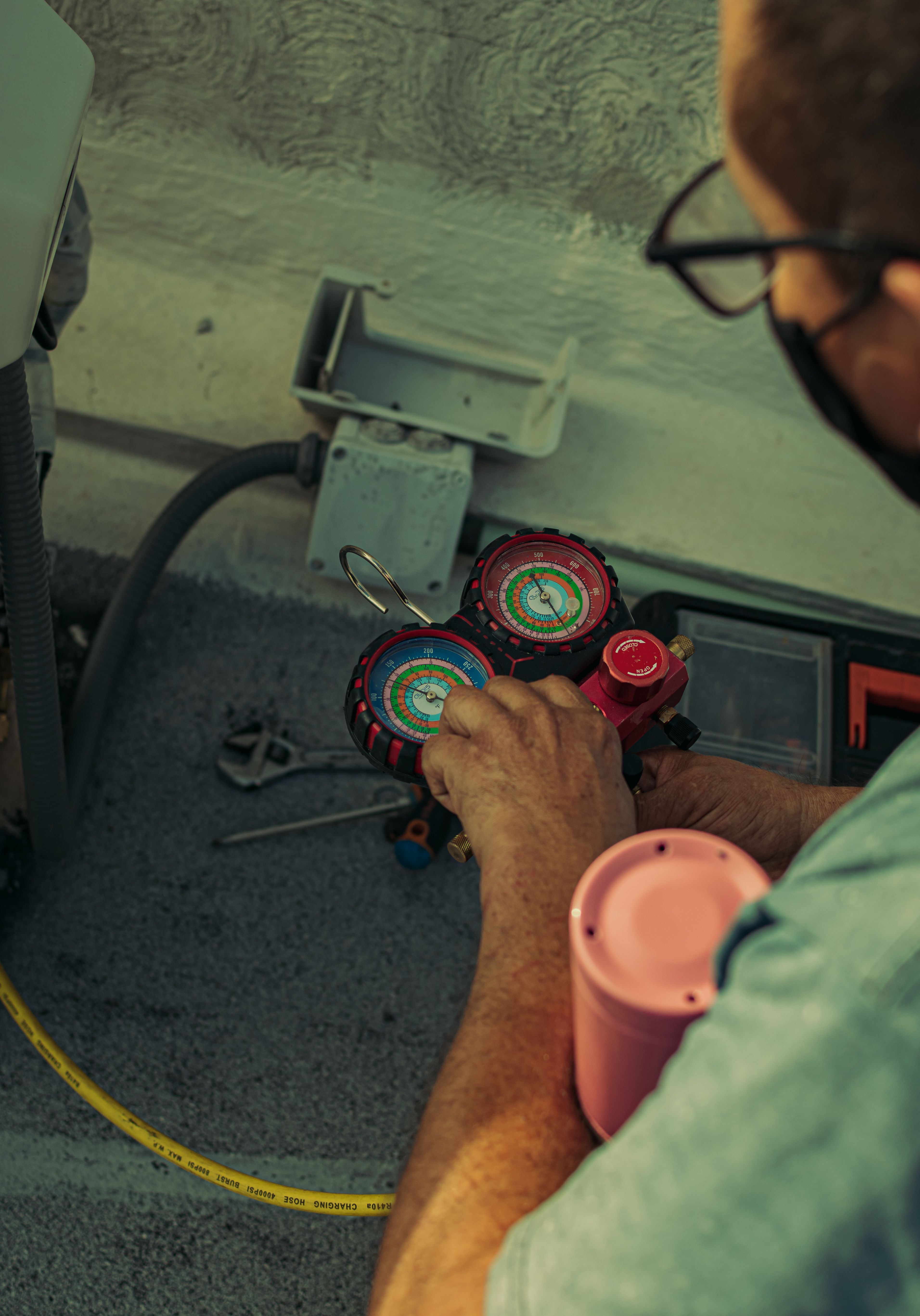 Technician gripping a red refrigerant manifold gauge set with a yellow charging hose while diagnosing an outdoor air conditioning unit beside its electrical disconnect box