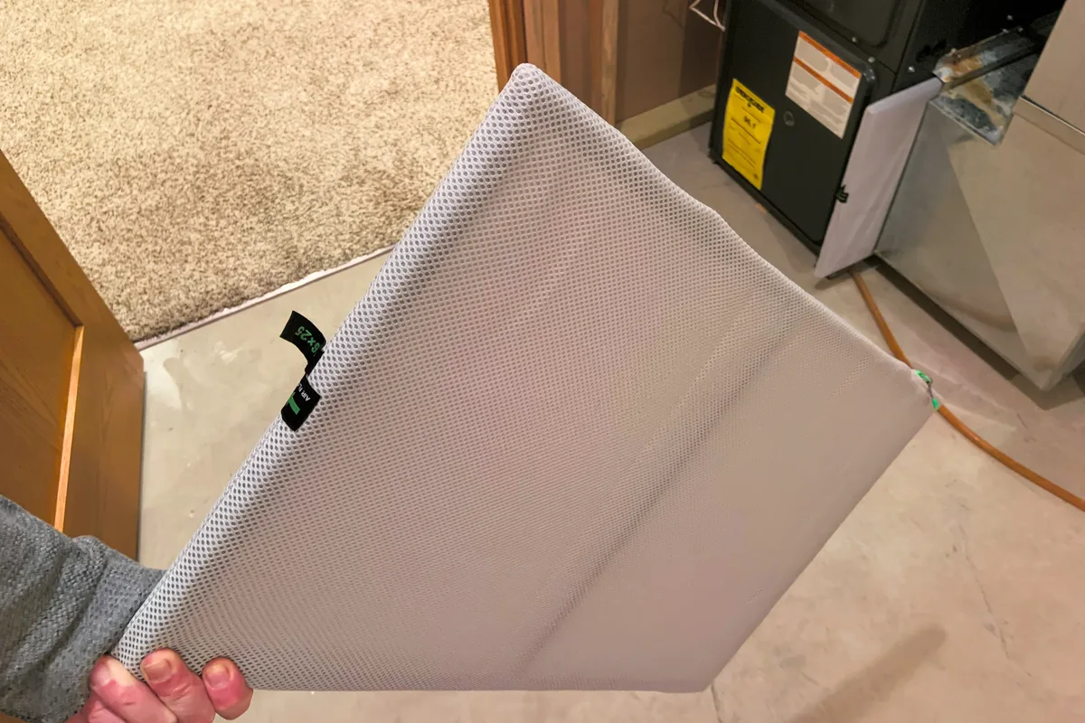 A homeowner holding a 16 by 25 inch furnace filter in front of a gas furnace in a home utility closet